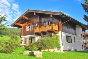 Ein zweistöckiges Holzhaus mit einem Balkon, umgeben von Grünflächen und blauem Himmel.