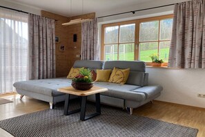 A modern living room with a grey sofa, a wooden coffee table, and a view of a green landscape through a large window. - Holiday apartment with 1 bedroom (Bischofswiesen)