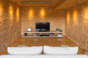 A cozy room with a stone wall, wooden floor, a flat-screen TV on a shelf, and a white sofa. - Olea Villas - Amazing villas by the sea (1) (Chania)