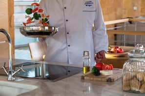 A chef holding a metal bowl with vegetables, a cutting board with tomatoes and cheese, and a jar of cookies on a kitchen counter. - Olea Villas - Amazing villas by the sea (1) (Chania)