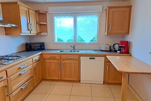 A compact kitchen with wooden cabinets, a dishwasher, a microwave, a gas stove, and a window with a view of greenery. - Holiday home in Hanvec for 5 people (Hanvec)
