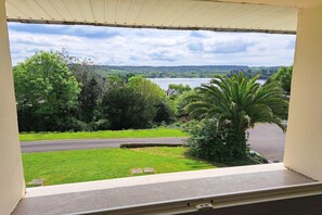 A window view of a residential area with a lawn, a palm tree, and a body of water in the distance. - Holiday home in Hanvec for 5 people (Hanvec)