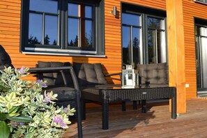 Um deck de madeira com móveis externos, incluindo um sofá e uma mesa, e uma janela com vista para árvores.