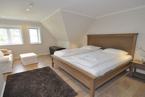 A bedroom with a large bed, a bedside table, a nightstand, a window, and a chair. - Old Rectory 5||St.Peter-Ording (St.Peter-Ording)