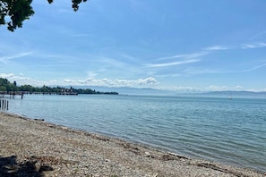 A pebbled beach with a pier extending into calm waters, a distant mountain range, and a clear sky. - Holiday apartment with 1 bedroom (Kressbronn)