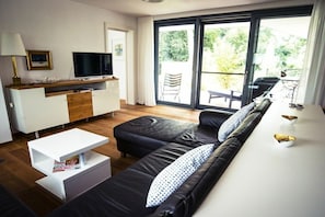A modern living room with a sofa, a coffee table, a TV, and a sliding glass door leading to an outdoor area. - Holiday apartment with 1 bedroom (Kressbronn)