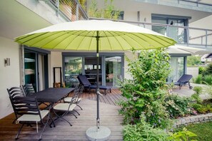 A patio with a table and chairs, a green umbrella, and a garden with plants and flowers. - Holiday apartment with 1 bedroom (Kressbronn)