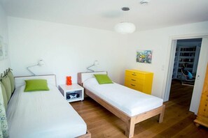 Een kamer met twee eenpersoonsbedden, een klein wit nachtkastje, een gele dressoir en een deur naar een gang.