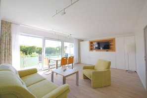 A modern living room with a sofa, armchair, and a dining table with chairs. A TV is mounted on the wall, and there is a sliding glass door leading to an outdoor area. - Seestern 9 Apartment House Seestern (Norderney)