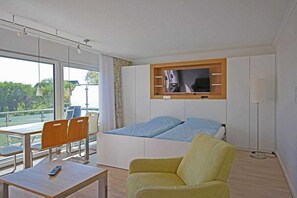 Habitación de hotel moderna con cama, televisor montado en un panel de madera, escritorio con silla y ventana con vista a follaje.