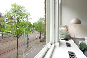 Ein Zimmer mit Fenster und Blick auf eine Straße, einem Bett mit Kopfteil und einer Stehlampe.