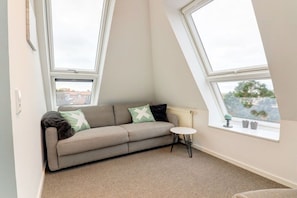 A room with a sofa, a small round table, and two windows with a view of buildings and trees. - Villa by the sea, upper deck 6 (Sankt Peter-Ording)