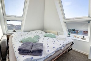 A small, triangular room with a bed, two windows, and a view of the city. - Villa by the sea, upper deck 6 (Sankt Peter-Ording)
