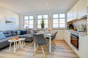 A modern kitchen with white cabinets, a dining area with a table and chairs, and a gray sofa with a fruit bowl. - Villa by the sea, upper deck 6 (Sankt Peter-Ording)