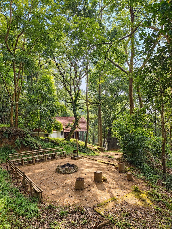 A forested area with a wooden deck, a fire pit, and a building with a red roof.