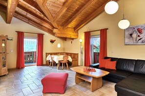 A cozy living room with a wooden ceiling, a black leather sofa, a red ottoman, a wooden coffee table, and a dining area with a table and chairs. - 6 Country House Farmer (Bodenmais)