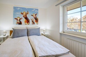 A double bed with a light-colored duvet, two bedside tables with lamps, a window with a view of a snowy landscape, and a painting of cows on the wall. - Holiday apartment with 1 bedroom (St. Peter-Ording)