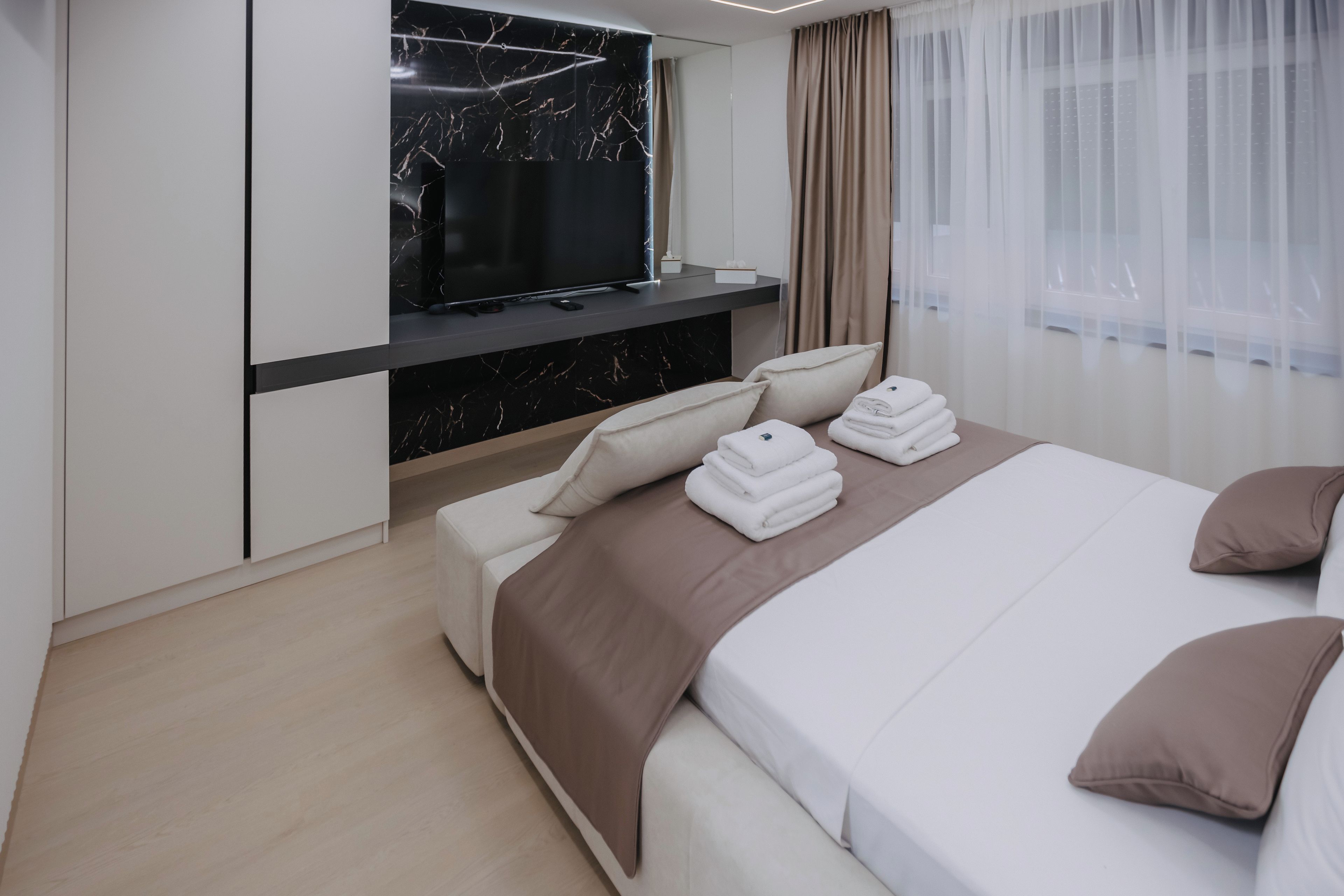 A modern hotel room with a flat-screen TV mounted above a marble fireplace, a bed with neatly folded towels, and a large window with sheer curtains.