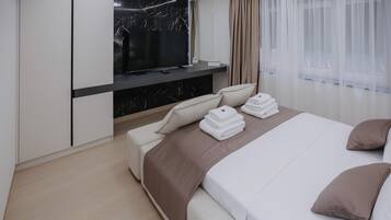 A modern hotel room with a flat-screen TV mounted above a marble fireplace, a bed with neatly folded towels, and a large window with sheer curtains.