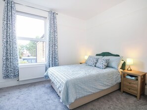 A bedroom with a bed, a nightstand with a lamp, and a window with curtains. - Fernleigh (Margate)