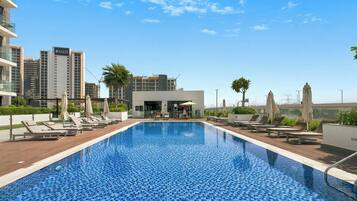 Una piscina sul tetto con lettini e ombrelloni, circondata da edifici moderni e un cielo sereno.
