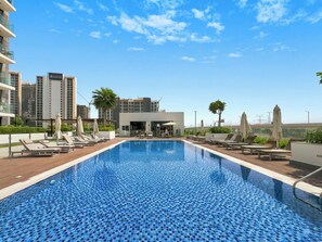 Piscina no terraço com espreguiçadeiras e guarda-sóis, cercada por edifícios modernos e um céu claro.