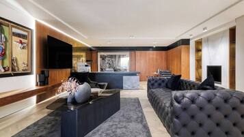 Uma sala de estar moderna com um sofá preto grande, uma mesa de centro de vidro e uma TV de tela plana fixada na parede.