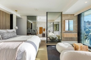 Quarto de hotel moderno com cama grande, área de estar, divisória de vidro e vista para a cidade.