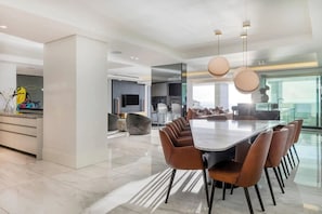 A modern dining area with a long table and multiple chairs, a kitchen in the background, and a large window providing a view. - Aurum Presidential Palace (Cape Town)