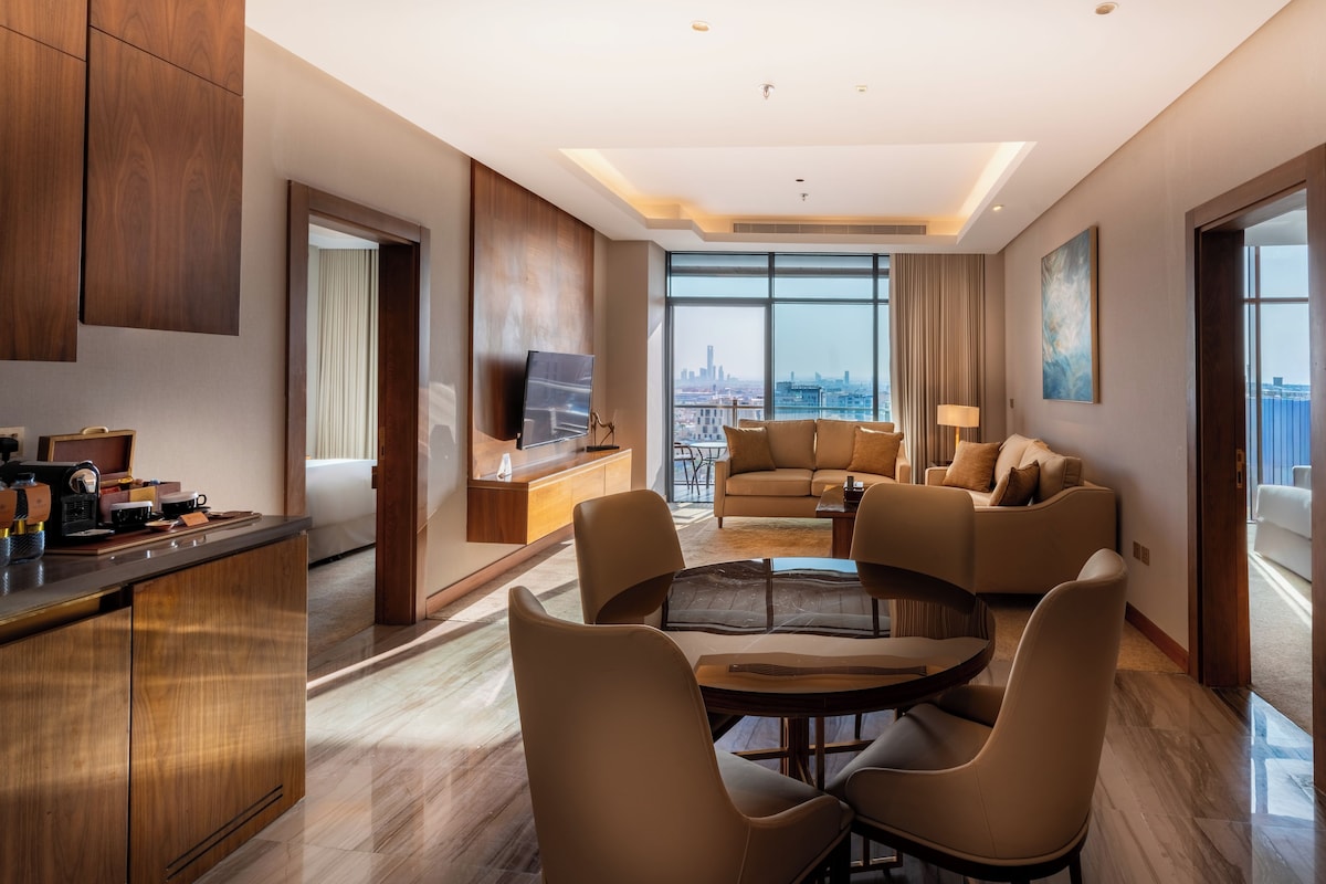 A modern hotel room with a city view, a dining area, and a living room with a sofa set.