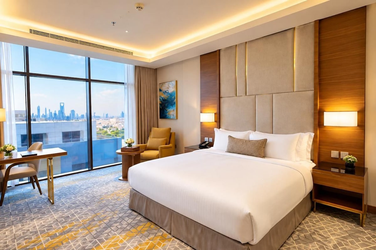 A hotel room with a large bed, a desk, a chair, and a view of a city skyline through large windows.