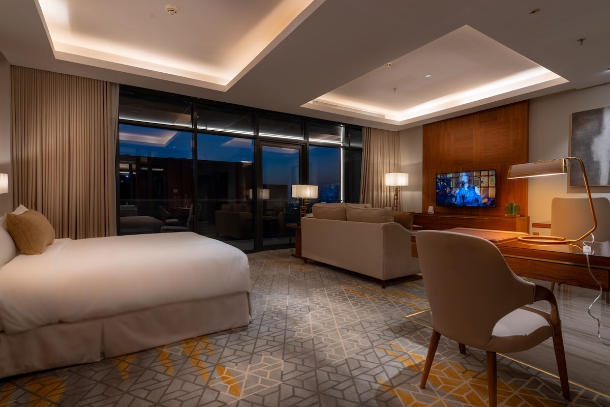 A hotel room with a bed, a sofa, a desk with a chair, a television, and a large window with a city view.