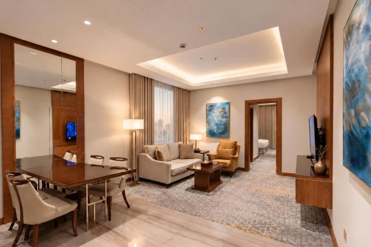 A hotel room with a dining table, sofa, and armchairs, a large mirror, and artwork on the walls.