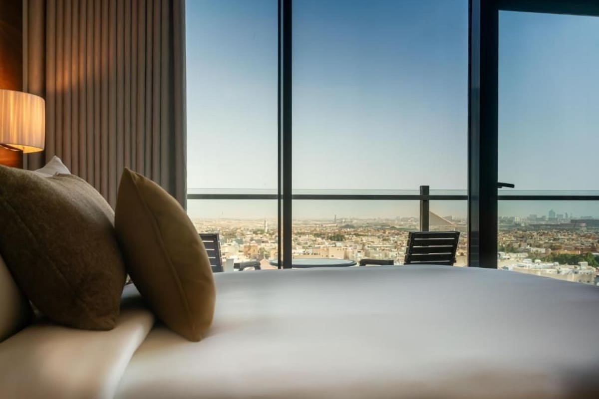 Ein Hotelzimmer mit einem großen Bett, Blick auf eine Stadtlandschaft durch raumhohe Fenster und einer Lampe auf der linken Seite.