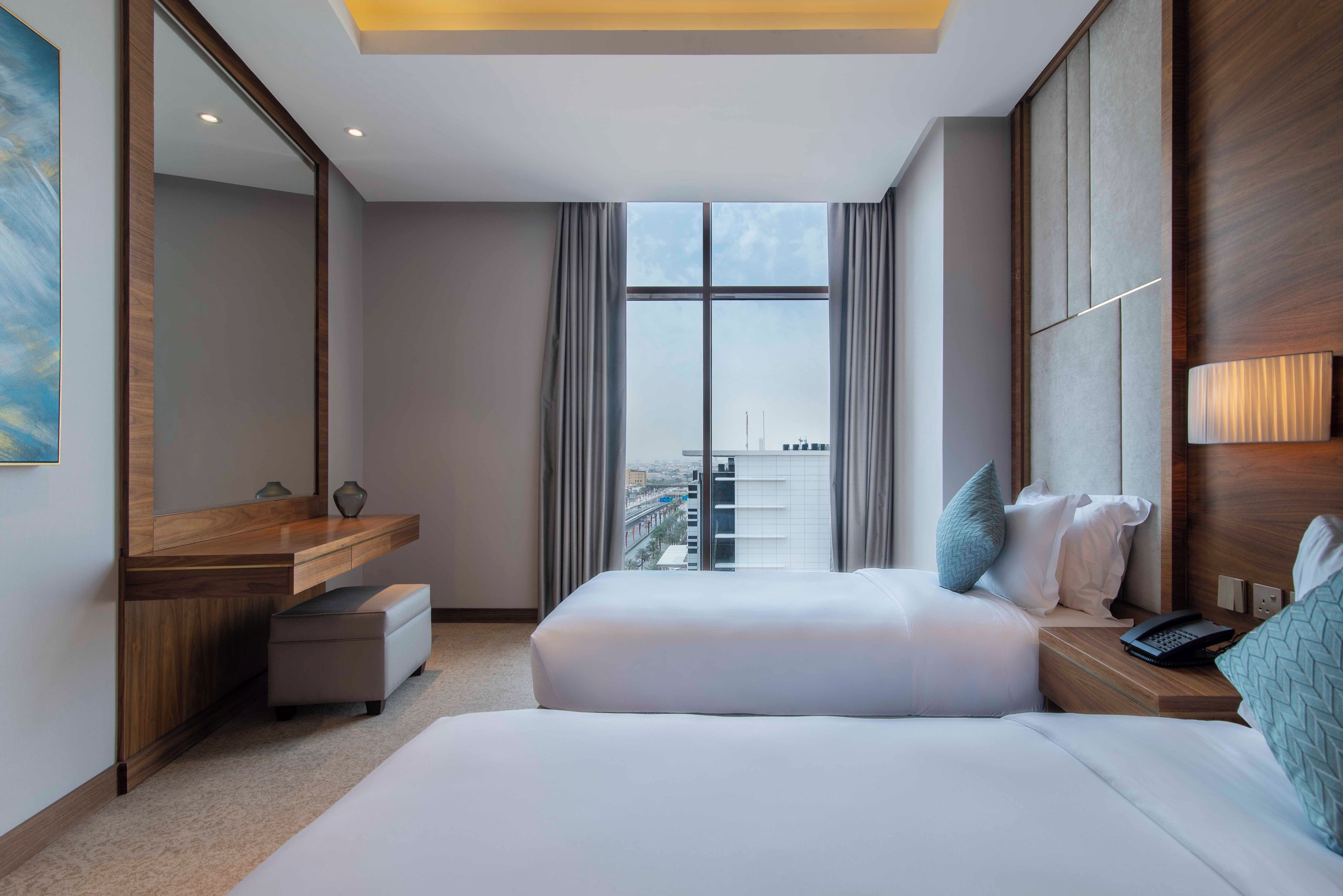 A hotel room with a large bed, a bedside table, a mirror, a window with a city view, and a small desk with a lamp.
