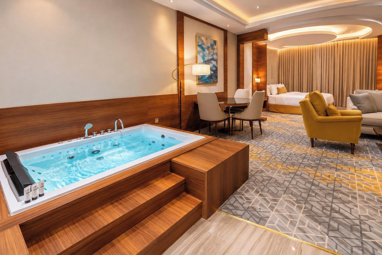 A hotel room with a large bathtub, a dining area with chairs, a sofa, and a patterned carpet.