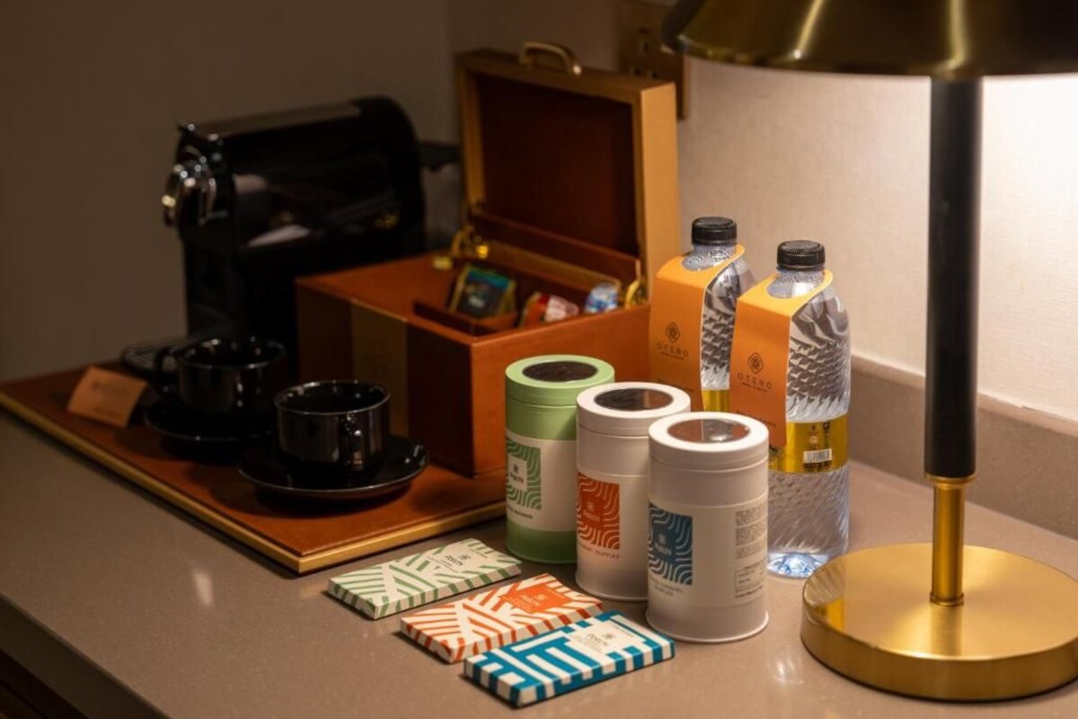 Ein Hotelzimmer mit Kaffeemaschine, einem Tablett mit Tassen und einer Schachtel, einer Lampe sowie Wasser- und Saftflaschen.