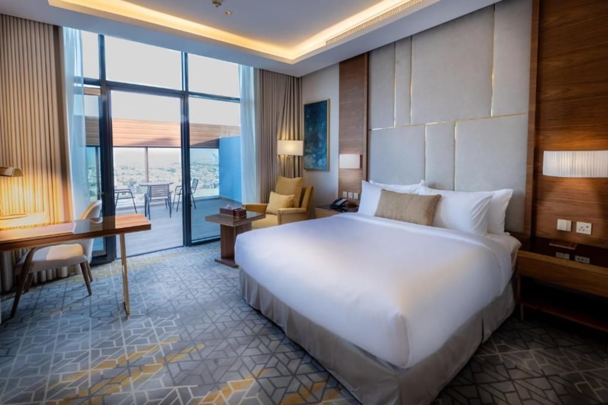 Ein Hotelzimmer mit einem großen Bett, einem Schreibtisch, einem Sessel, einem kleinen Tisch und Blick durch die Glastür.