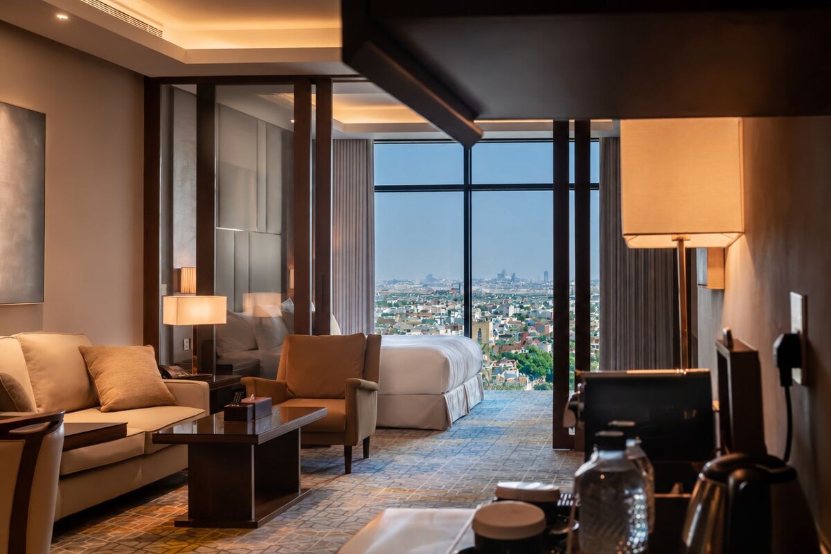 A modern hotel room with a city view, a sofa, a chair, a coffee table, and a desk.