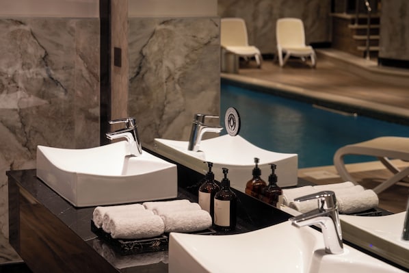 A bathroom with two sinks, a mirror, and a pool area in the background. - Otero Hotel and Suites - Riyadh (Riyadh)