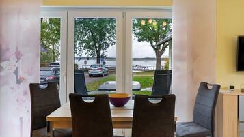 Een eetgedeelte met een tafel, stoelen, een schaal en uitzicht op een waterpartij met auto’s en bomen.