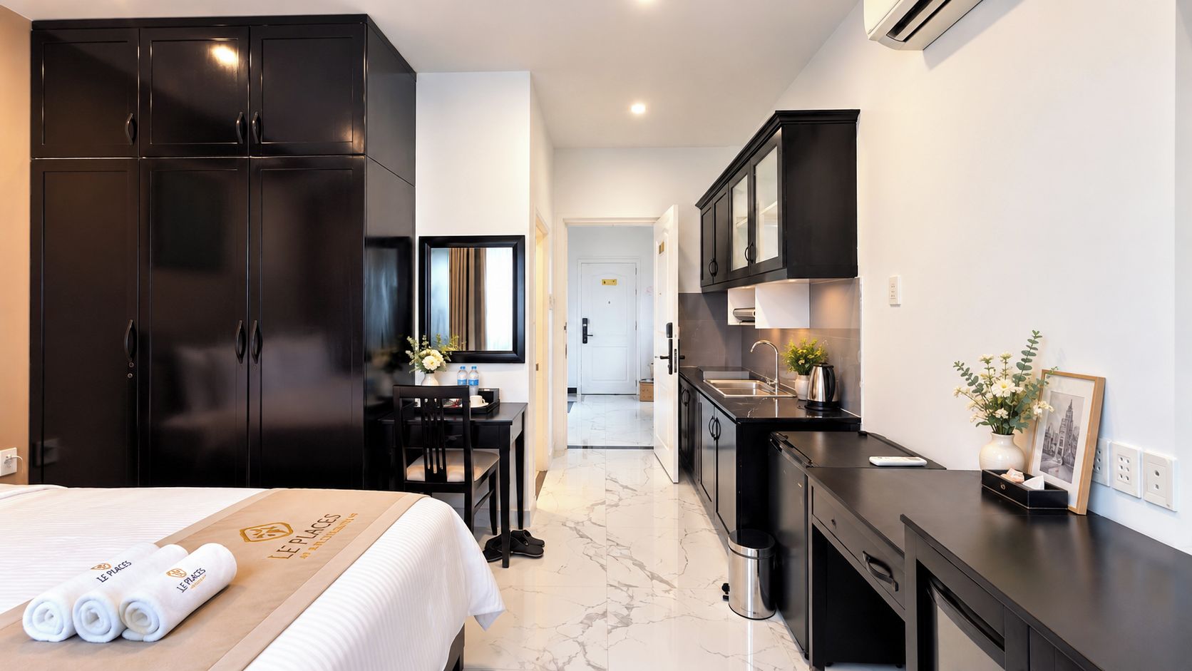 Une chambre d’hôtel avec un lit, un bureau, une chaise, une armoire, un coin cuisine et une salle de bain que l’on aperçoit à l’arrière-plan.