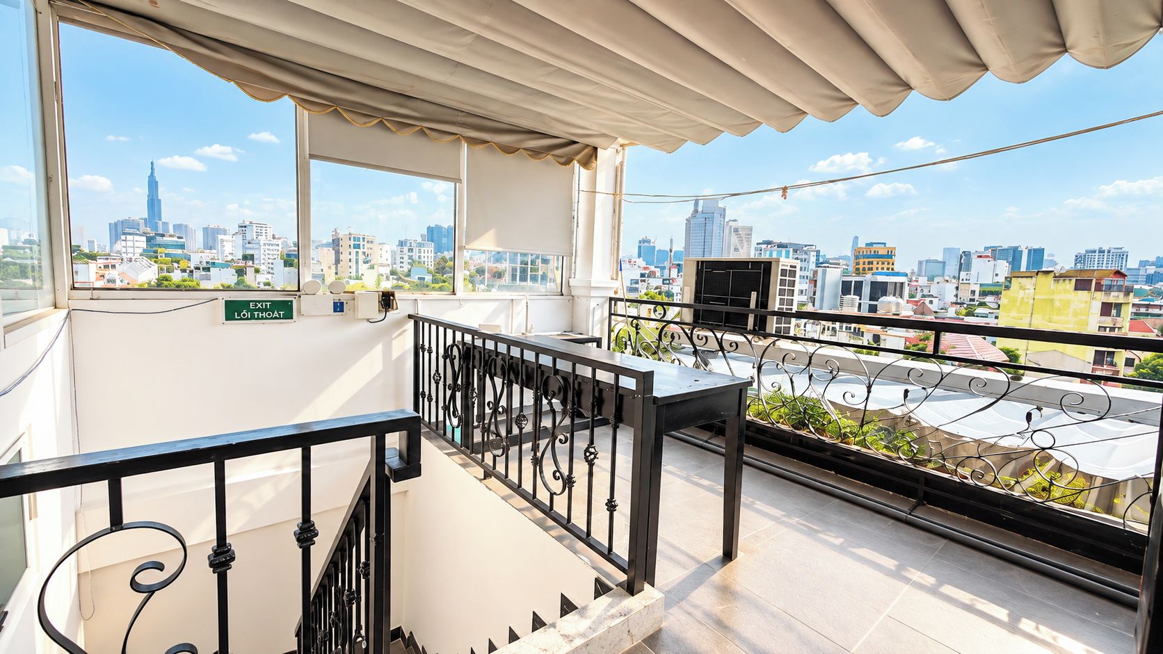 Un balcon doté d’une balustrade en métal, d’une table et offrant une vue sur le paysage urbain, notamment sur un immeuble de grande hauteur.