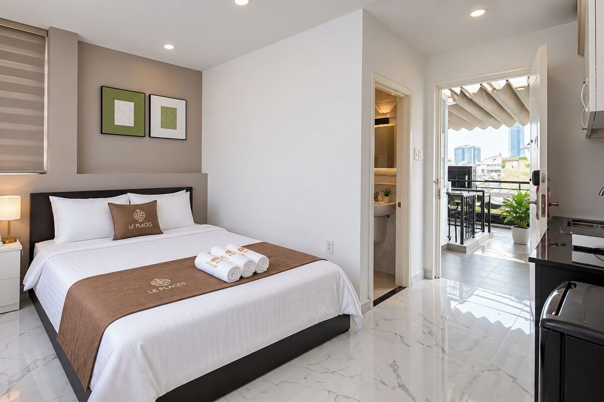 A hotel room with a bed, a nightstand with a lamp, a window with blinds, and a view of the city through the door.