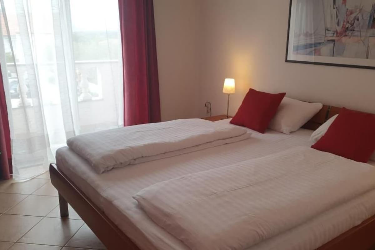 Ein Doppelbett mit weißer Bettwäsche und roten Kissen, ein Nachttisch mit Lampe und ein Fenster mit durchsichtigen Vorhängen.