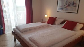 Ein Doppelbett mit weißer Bettwäsche und roten Kissen, ein Nachttisch mit Lampe und ein Fenster mit durchsichtigen Vorhängen.