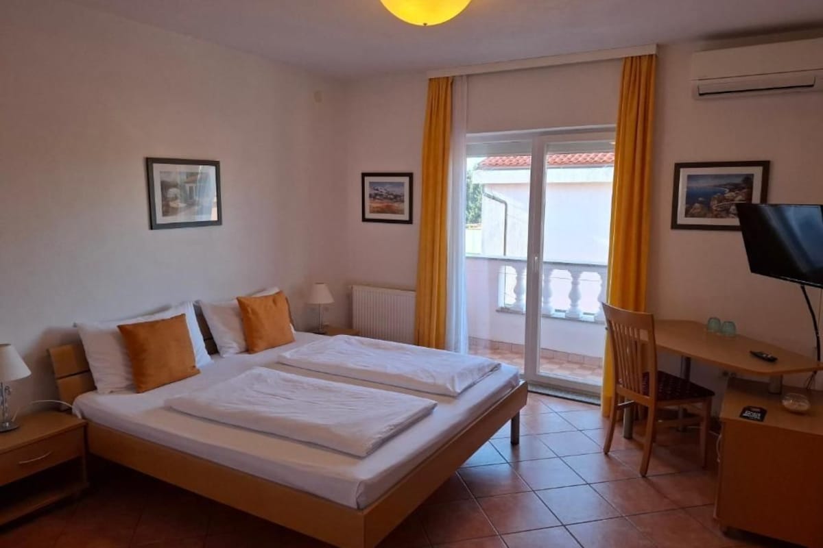 Ein Hotelzimmer mit einem Doppelbett, einem Schreibtisch mit Stuhl, einem Fernseher und einem Balkon mit gelben Vorhängen.