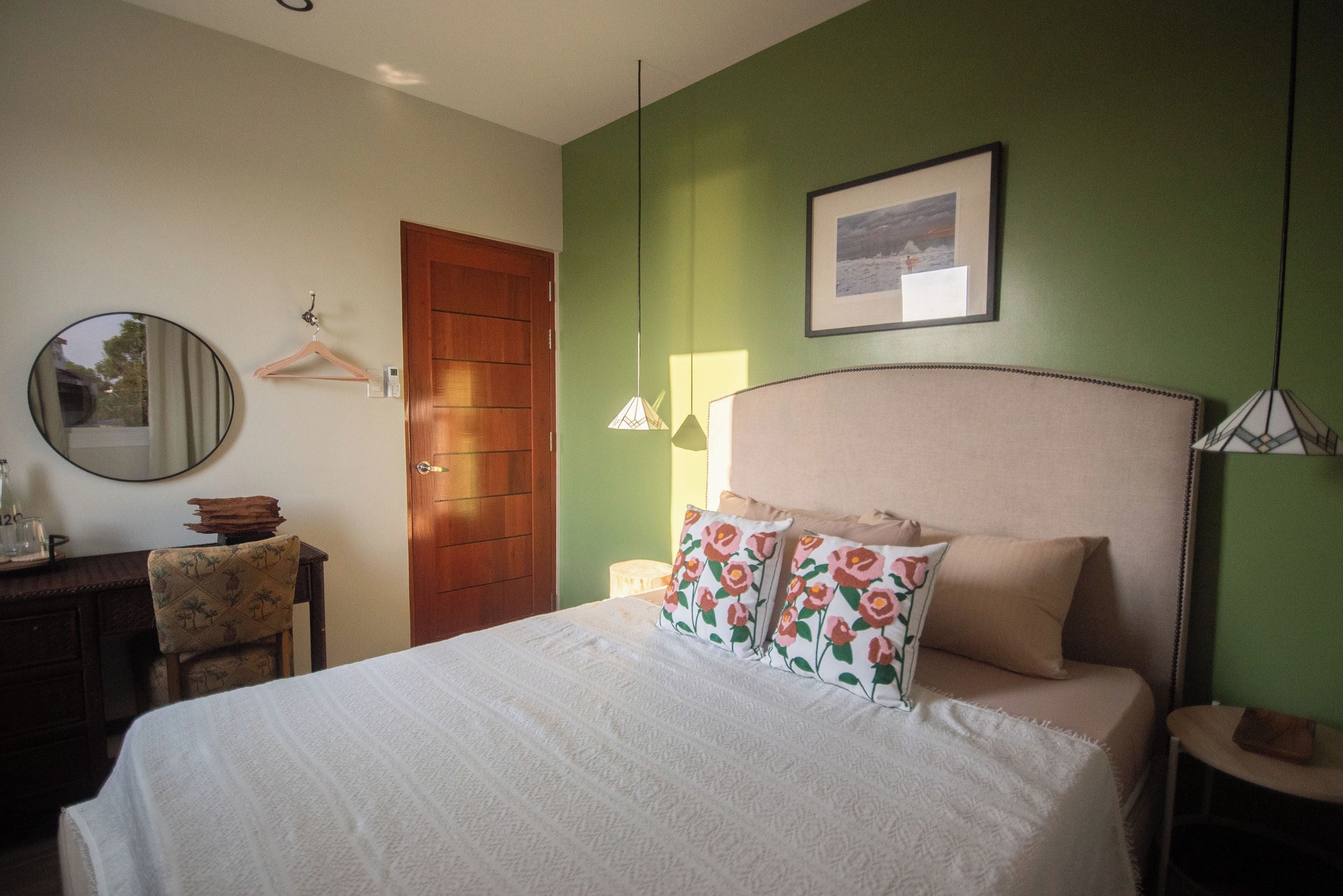 A bedroom with a bed, a wooden door, a mirror, a framed picture on the wall, and a small table with a chair.