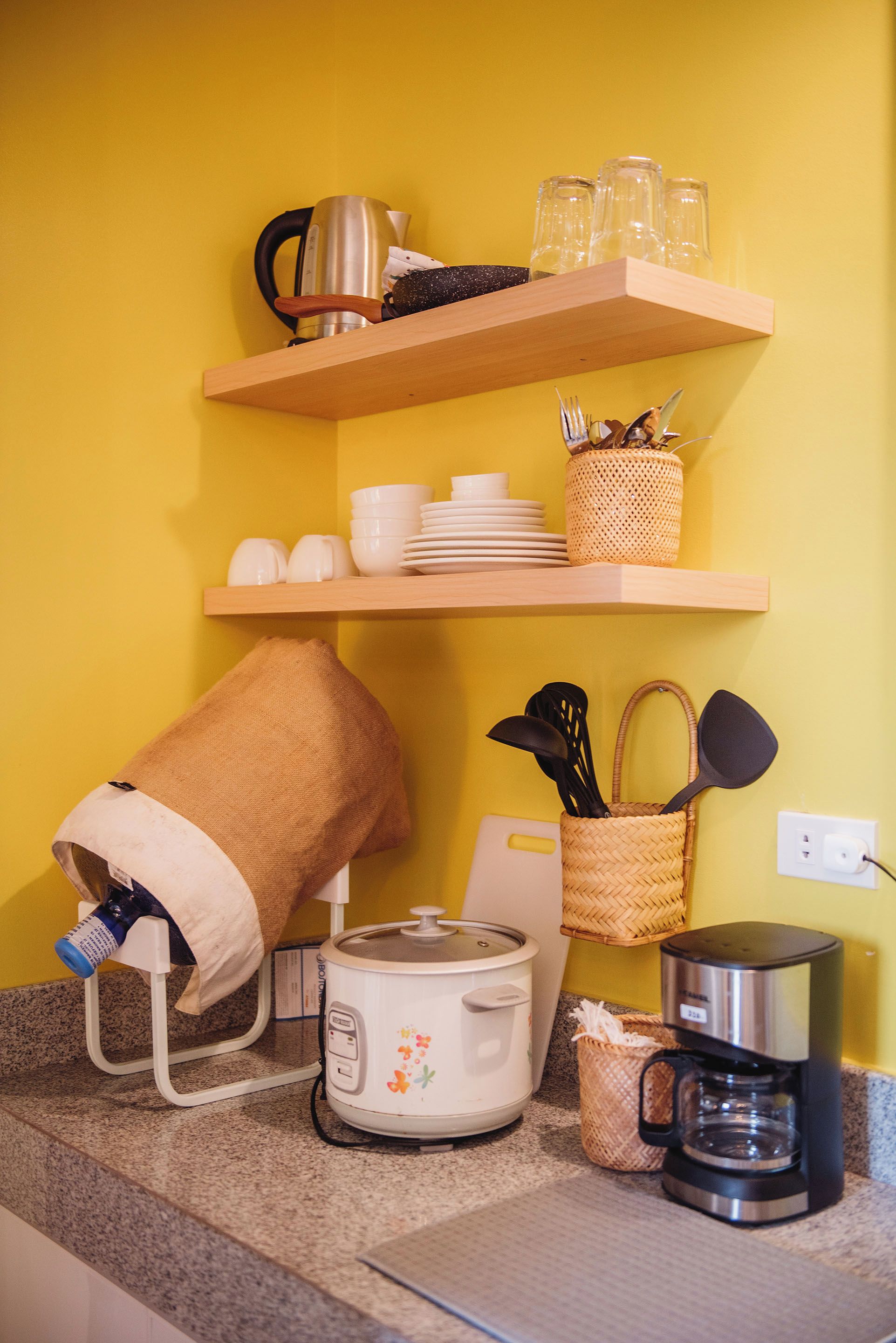 A kitchen with a yellow wall, wooden shelves, a kettle, a coffee maker, a rice cooker, and various kitchen utensils.