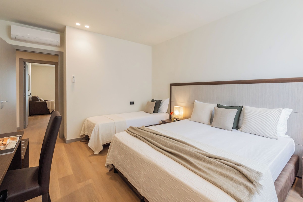 A hotel room with a large bed, two bedside tables, a desk with a chair, and a door leading to another room.